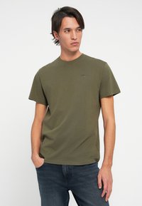 Mladý muž oblečený v olivově zeleném tričku Tommy Jeans a tmavě modrých džínách, stojící s jednou rukou v kapse a dívající se do strany.
