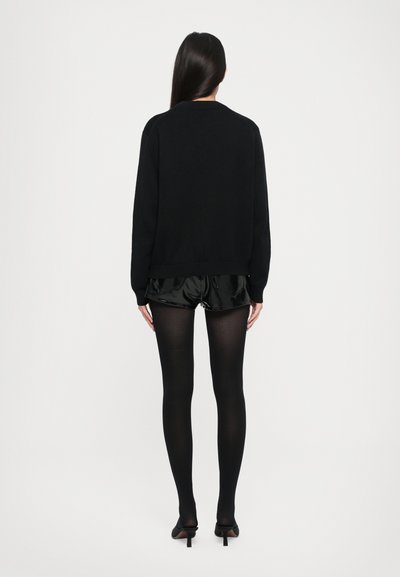 Svart strikket genser, svarte blanke shorts under, svarte strømpebukser og svarte hæler, vist bakfra mot en enkel bakgrunn.