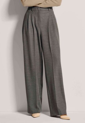 Femme portant un pantalon gris taille haute à jambes larges avec plis, des chaussures pointues beiges et un pull beige, bras croisés.