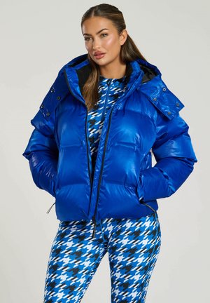 Blauwe puffjack met een glanzende afwerking, rits aan de voorkant, oversized capuchon en drukknoopjes op de schouders, gecombineerd met leggings met houndstooth-print.