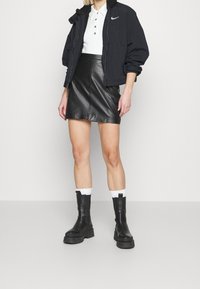 Svart bomberjacka med shearling-krage, vit polo-tröja, läder mini kjol och ankelstövlar med kraftiga sulor. Vita crew-strumpor synliga.