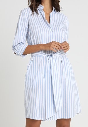 Femme portant une robe chemise à manches longues, longueur genoux, avec des rayures verticales bleu clair et blanc, cintrée à la taille avec une ceinture assortie.