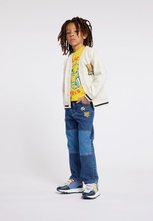 Gilet blanc avec des bordures bleu marine, t-shirt graphique jaune, jeans en denim bleu avec des patches, et baskets multicolores.