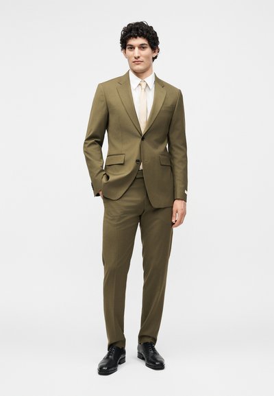 Άνδρας με σγουρά μαλλιά που φοράει ελαιί πράσινο κοστούμι, λευκό πουκάμισο, μπεζ γραβάτα και μαύρα επίσημα παπούτσια, στέκεται με το ένα χέρι στην τσέπη.