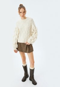 Kremowy sweter dziany z frędzlami na rękawach, wzorzysta mini spódnica oraz brązowe skórzane kozaki do kolan z klamrami i kontrastującymi skarpetkami.