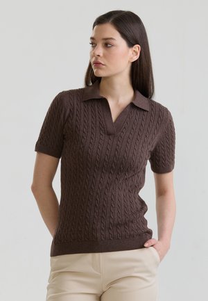 Junge Frau mit langen dunklen Haaren, die ein braunes, kurzärmeliges, kabelgestricktes Poloshirt und hellbeige Hosen trägt, Hände in den Taschen.