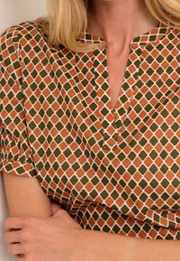 Haut à manches courtes présentant un motif géométrique dans des tons terreux de vert et d'orange sur un fond crème. Encolure en V avec manches froncées.
