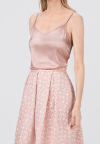 Žena v světle růžovém saténovém topu na tenkých ramínkách a v sukně s vysokým pasem a jemným květinovým vzorem.