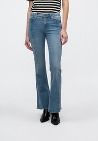 Verblasste blaue Denim-Flared-Jeans mit mittelhohem Bund, ausgestattet mit Fronttaschen und einem Knopfverschluss, kombiniert mit schwarzen, spitz zulaufenden Stiefeletten.