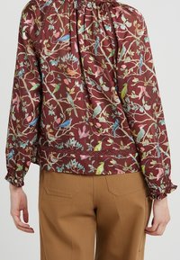 Blouse florale bordeaux avec des motifs d'oiseaux multicolores. Elle présente un col froncé, des poignets élastiques et un détail en dentelle décoratif à l'ourlet.