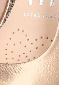 Plantilla de piel nude con pequeñas perforaciones, detalles metálicos en los bordes y texto plateado que dice "TOTAL FLEX". Textura suave y lisa.