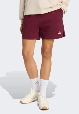 Bordowe sportowe szorty z elastycznym paskiem w talii, z małym różowym logo na obszyciu, zestawione z białymi skarpetkami i jasnymi trampkami.