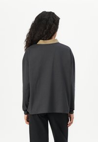 Kvinne med langt krøllete hår som står med ryggen til kameraet iført en mørk langermet skjorte med beige krage og svarte bukser.
