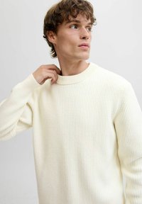 Maglione a coste di colore crema con collo alto e maniche lunghe, caratterizzato da una texture morbida e una vestibilità rilassata.