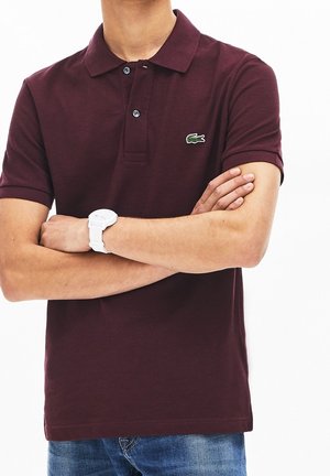 Mand iført en burgunder polo med krokodillelogo, blå jeans og hvid armbåndsur, armene krydset mod hvid baggrund.
