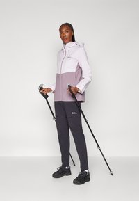 Veste imperméable légère en lilas et lavande avec un design bicolore, associée à un pantalon de randonnée noir et des bâtons de trekking.