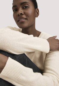 Cremefarbener, strukturierter Pullover mit gerippten Bündchen, getragen über dunklen Jeans. Der Fokus liegt auf dem weichen Material und der lässigen Passform.