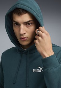Sweat-shirt à capuche teal en tissu doux, avec un logo Puma blanc sur la poitrine et un cordon de serrage pour l'ajustement. Doigt orné d'une bague.