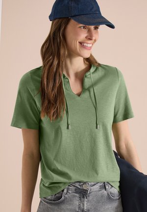 Smilende kvinde iført en grøn hættetrøje med korte ærmer, lysegrå jeans og en mørkeblå baseballkasket, der holder en mørk jakke ved sin side.