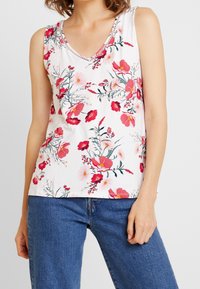 Donna con top bianco senza maniche con stampa floreale rosa e rossa e jeans di denim blu, in piedi contro uno sfondo chiaro e semplice.