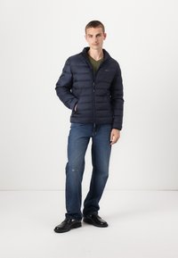 Marineblauwe gewatteerde jas met een ritssluiting aan de voorkant, hoge kraag en elastische manchetten, gecombineerd met blauwe jeans en zwarte schoenen, tegen een witte achtergrond.