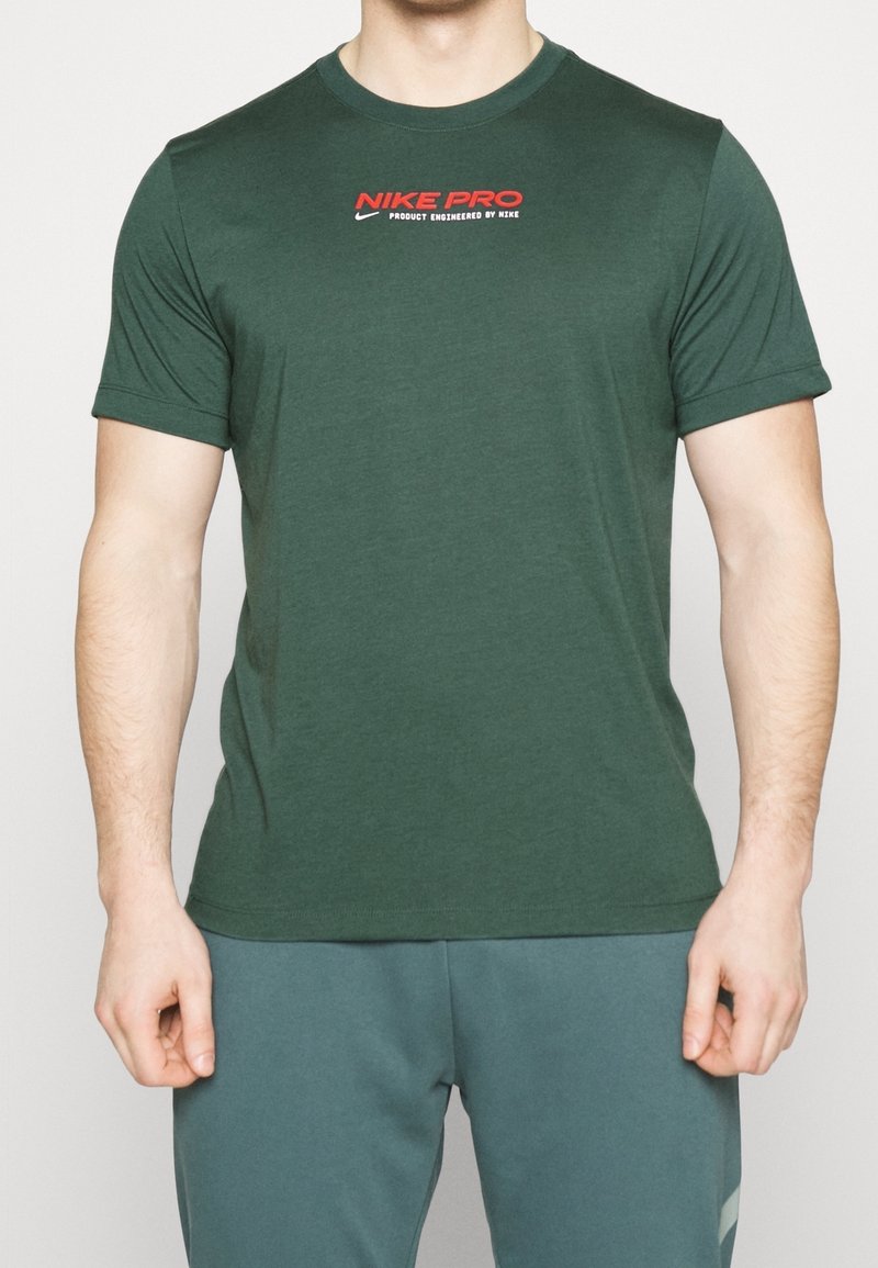 Homme portant un t-shirt à manches courtes Nike Pro vert foncé avec un logo rouge et un pantalon de sport vert foncé, debout devant un fond uni.