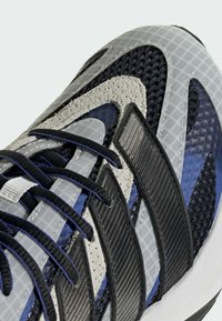 Gros plan sur une chaussure de sport bleue, noire et grise montrant un tissu en maille, des détails en daim, trois bandes noires et des lacets noirs.
