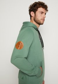 Grüne Kapuzenjacke mit einer Vorderentasche, ausgestattet mit einem braunen Lederpatch am linken Ärmel und schwarzen Kordeln. Weicher Stoff mit gerippten Bündchen.