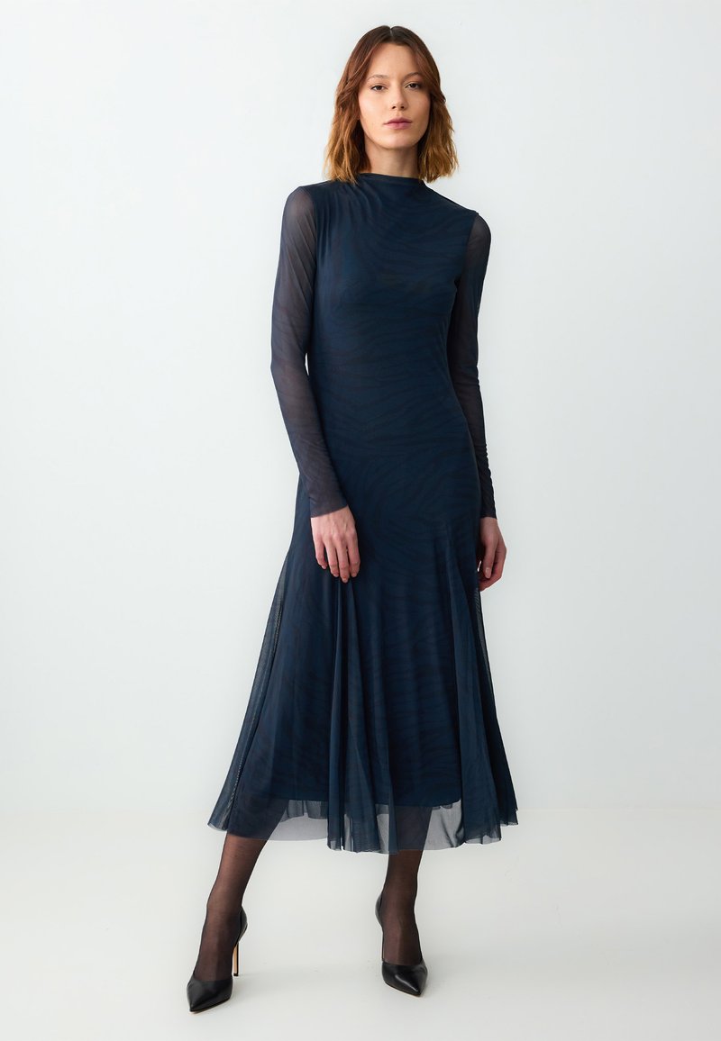 Abito lungo navy a maniche lunghe con gonna fluida, caratterizzato da un motivo testurizzato, maniche trasparenti e scollatura alta. Indossato con tacchi neri.