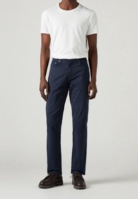 Chino in cotone blu navy con vestibilità dritta, dotato di cinque tasche, pattina con zip e chiusura a bottone. Abbinato a una maglietta bianca a collo rotondo.