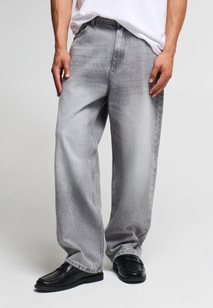 Uomo che indossa jeans larghi grigio chiaro, mocassini neri e una maglietta bianca infilata, in piedi davanti a uno sfondo chiaro e uniforme.