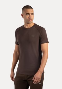 Camiseta marrón de manga corta hecha de tela suave, con cuello redondo y una pequeña etiqueta en el pecho. Textura suave y diseño ajustado.
