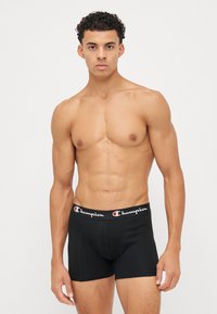 Black cotton boxer briefs with a stretchy waistband featuring the "Champion" logo in white, red, and black. Fitted design, smooth texture.