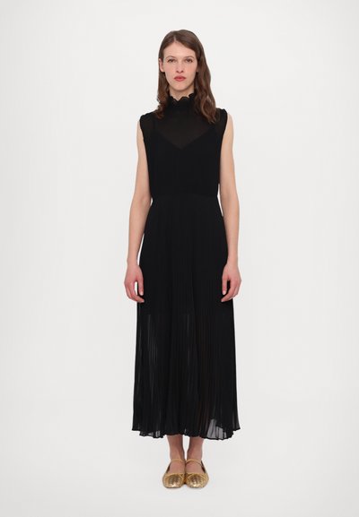 Schwarzes plissiertes Kleid mit ärmellosem Design, hohem Kragen und transparentem Oberteil. Kombiniert mit goldenen Slippern. Einfarbig weißer Hintergrund.