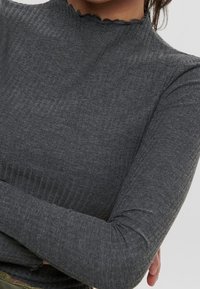 Nahaufnahme einer Person, die ein enganliegendes, geripptes, dunkelgraues Longsleeve mit einem gewellten hohen Halskragen trägt und die Arme überkreuzt.
