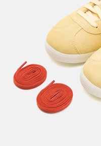 Zapatillas de ante amarillas con suelas de goma blanca, acompañadas de dos cordones de algodón rojos enrollados ordenadamente a su lado.