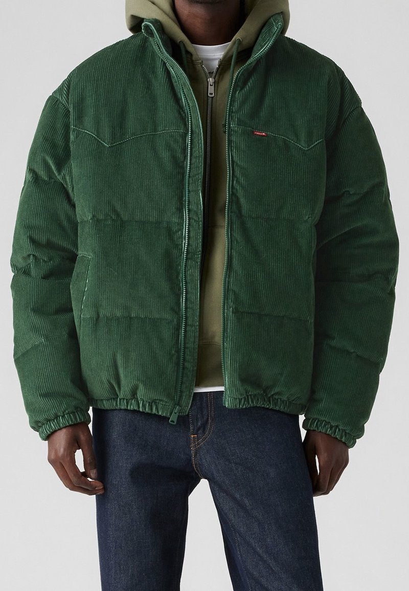 Zielona kurtka pikowana z aksamitu sztruksowego o pikowanej fakturze, z zamkiem błyskawicznym, ściągaczami na mankietach i u dołu. Noszona na szarej bluzie z kapturem i ciemnych dżinsach.