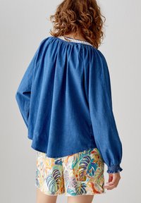 Top blu lungo con scollo arricciato e polsini elasticizzati, abbinato a pantaloni corti colorati e fantasiosi con motivi di foglie tropicali.