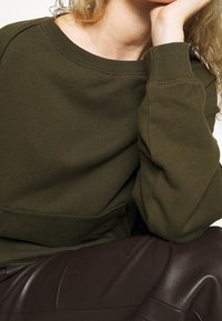 Sudadera verde oliva con cuello y puños acanalados, que presenta un corte relajado y una textura suave, combinada con pantalones de piel sintética marrón.