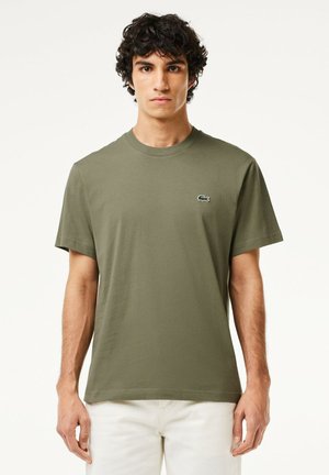T-shirt en coton vert olive avec un col rond. Il a des manches courtes et un petit logo vert et blanc sur la partie supérieure gauche de la poitrine.