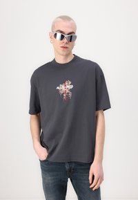 Camiseta de algodón gris con un diseño floral y el texto "ALL SAINTS" en el centro. Modelo usando gafas de sol reflectantes y jeans azules.