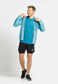 Lichtgewicht, turquoise hardloopjack over een gestreept blauw shirt, gecombineerd met zwarte shorts en sportschoenen. Soepele textuur, functioneel ontwerp.