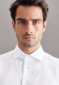 Witte overhemd met een klassieke kraag, knoopdetails en een gladde stoftextuur, gedragen door een model tegen een neutrale achtergrond.