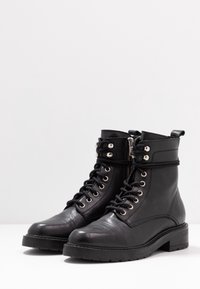 Botas de tobillo de cuero negro con puntera redonda, cordones en la parte delantera, acentos de metal plateado y cremallera lateral. Parte superior texturizada y suela gruesa.