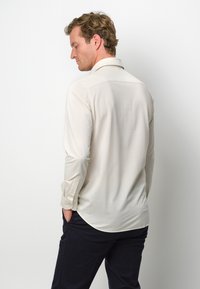 Camicia a maniche lunghe beige chiaro con colletto, orlo arrotondato e polsini bottonati, realizzata in tessuto liscio, abbinata a pantaloni blu scuro.