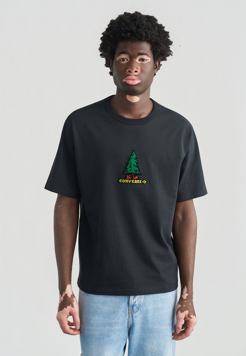 Μαύρο βαμβακερό t-shirt με πράσινο κεντημένο λογότυπο δέντρου και την επιγραφή "CONVERSE" σε κίτρινο από κάτω. Κανονική εφαρμογή με κοντά μανίκια. Ανοιχτό μπλε παντελόνι τζιν.