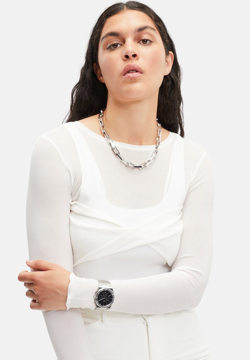 Hvid langærmet top med et lagdelt design. Modellen bærer en sølv kæde og et ur med sort urskive og sølvrem.