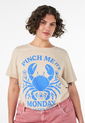Kvinde med kort krøllet hår iført beige T-shirt med blå krabbeprint og teksten "Nip mig, det er mandag", kombineret med lyserøde bukser.