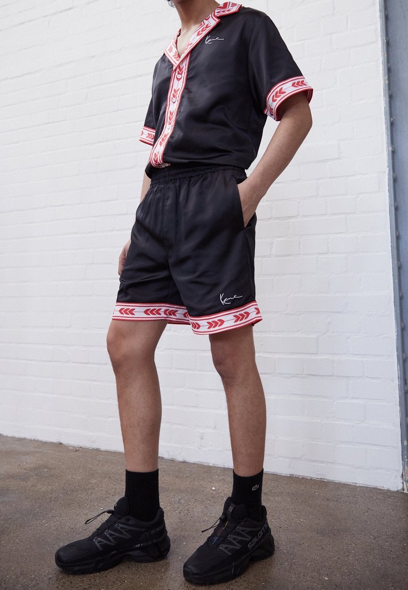 Chemise noire à manches courtes avec des bordures à motifs rouge et blanc, assortie de shorts noirs avec des bordures rouges. Portée avec des chaussures de sport noires.