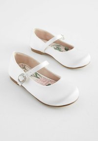 Chaussures Mary Jane en satin blanc avec un bout rond, une bride de cheville et une boucle décorative en argent. Doublure intérieure à motif floral. Semelles plates.